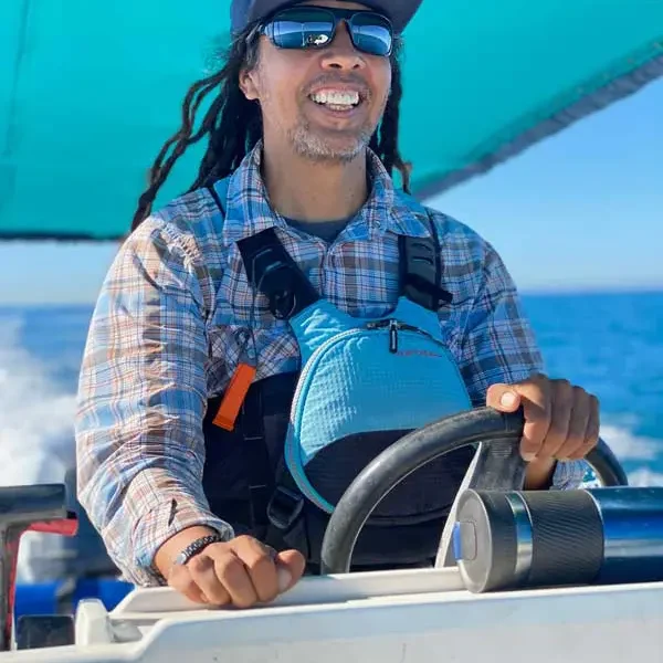 Person wearing sunglasses, a cap, and a plaid shirt steers a boat on the ocean under a teal canopy, smiling in bright daylight.